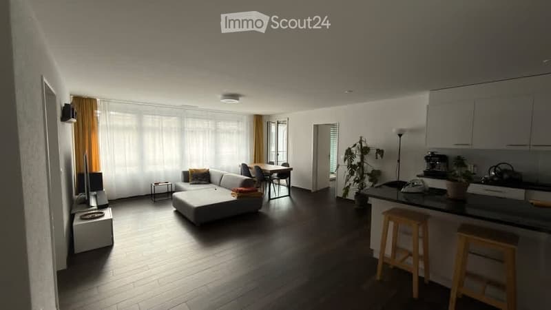Open-plan living and dining area with a grey sectional sofa, coffee table, dining table with chairs, kitchen island with stools, and a potted plant. The space features large windows with sheer curtains and wooden flooring.