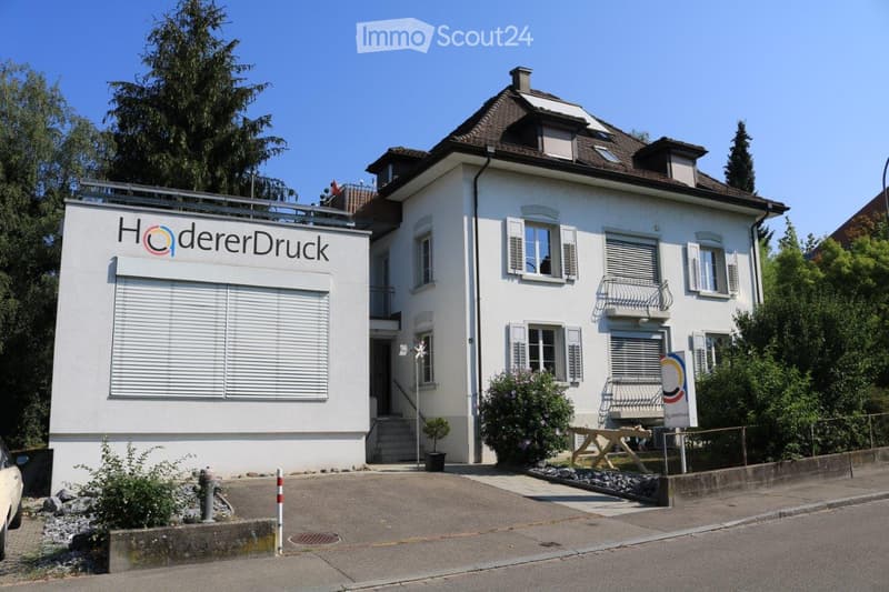 Two-story white building with a brown roof, multiple windows, and a sign reading "HödererDruck" on the side.