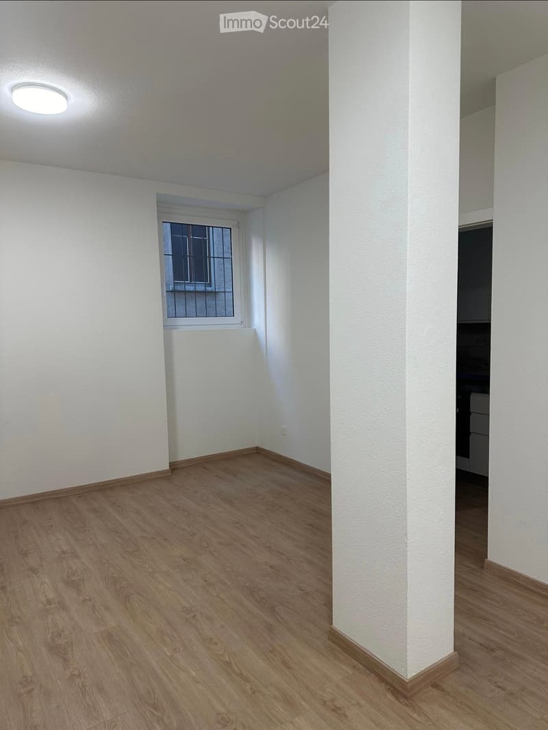Empty room with wooden flooring, white walls, and a single window with bars. The room has a ceiling light fixture and a visible doorway leading to another space.
