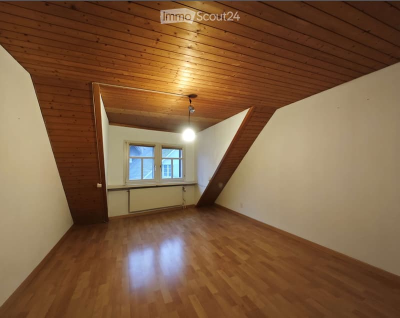 Empty room with wooden flooring, white walls, and a wooden slanted ceiling. Features a window with white frames and a radiator below it.