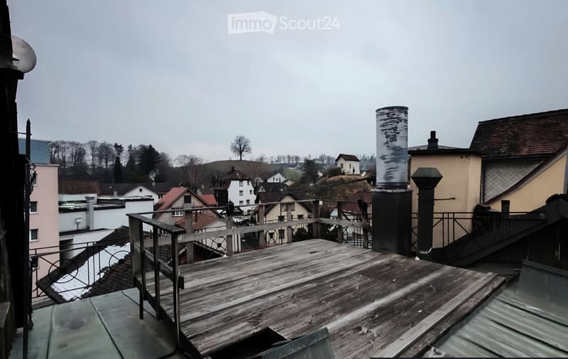 Roof terrace with wooden decking, metal railing, and view of surrounding houses and landscape.