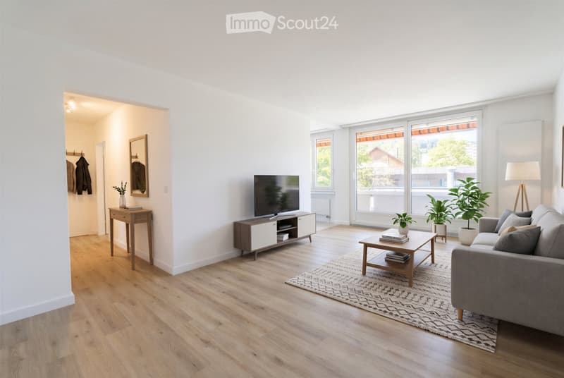 Bright living room with wooden flooring, large windows, grey sofa, coffee table, TV stand, side table, floor lamp, potted plants, and a coat rack in the adjacent hallway.