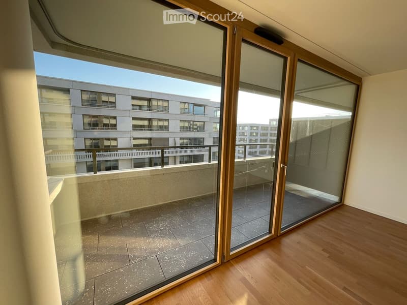 Empty room with wooden flooring and large glass sliding doors leading to a tiled balcony area with a view of a modern building.
