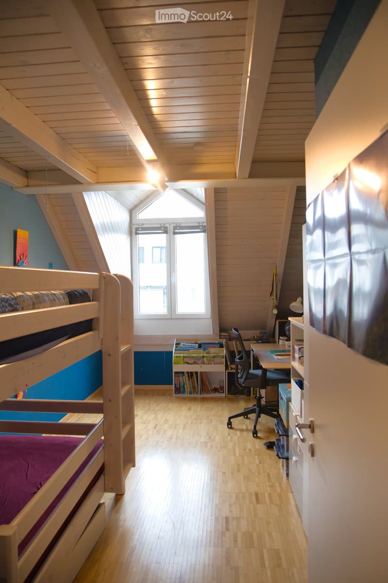 Bright attic bedroom with wooden bunk bed, desk, bookshelf, and a large window letting in natural light.