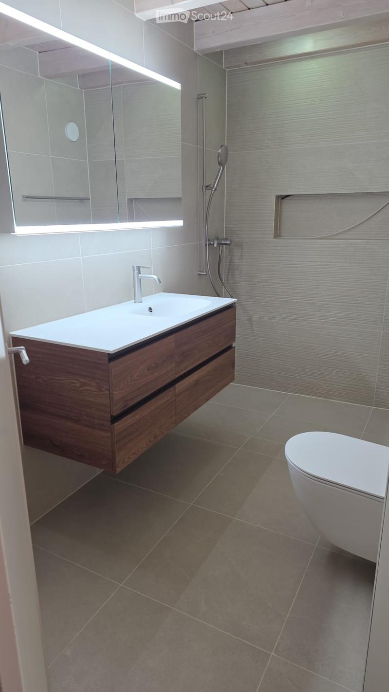 Modern bathroom with a wooden vanity, white sink, chrome faucet, and a white toilet. The shower area features a glass enclosure with a modern showerhead.