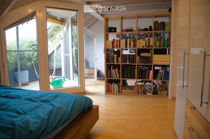 Bright bedroom with wooden floors, large windows, a bed with blue bedding, a bookshelf filled with books, and a chair.