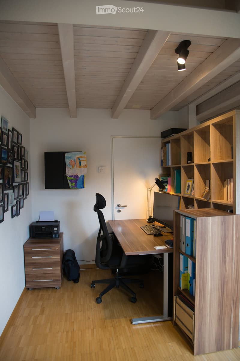 Modern office space with wooden flooring, slanted wooden ceiling, desk with ergonomic chair, bookshelf, printer, and framed artwork on the walls.