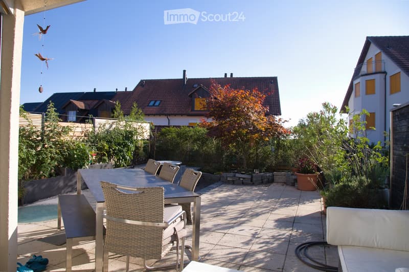 A sunny outdoor patio with a table, chairs, and a sofa, surrounded by greenery and a small swimming pool.