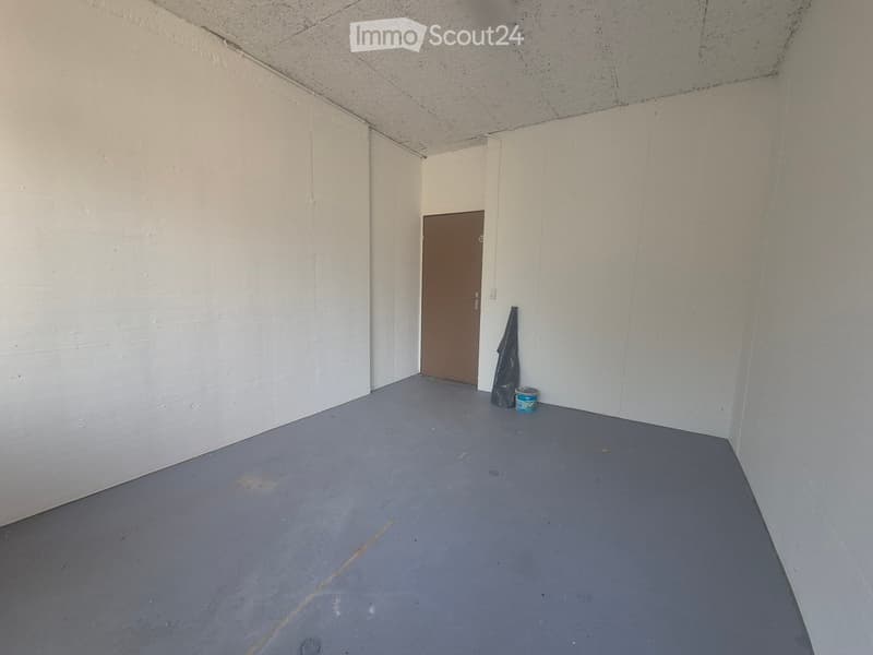 Empty room with white walls, grey concrete floor, and a single brown door leading to another space. A plastic bag and a small container are placed near the door.