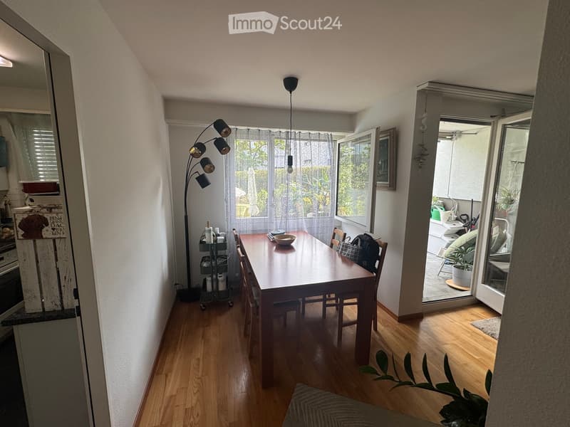 Dining area with wooden floor, dining table, chairs, floor lamp, and open sliding doors leading to a balcony or terrace with outdoor furniture.