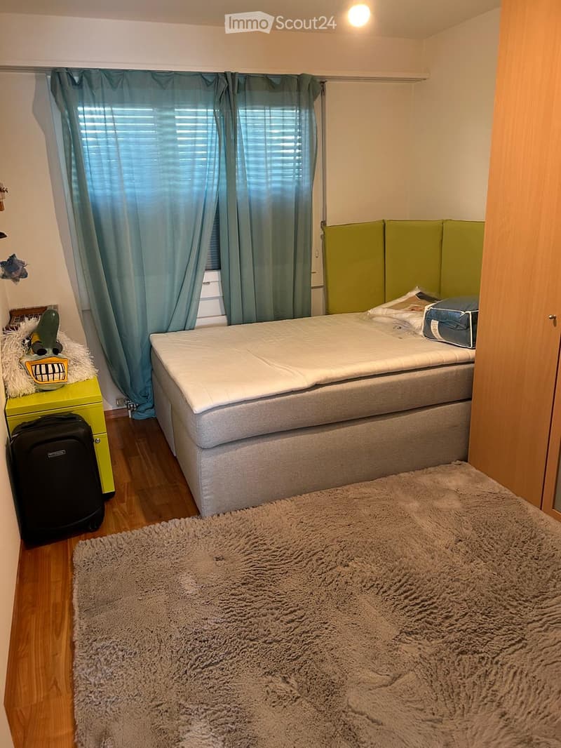 Cozy bedroom with a single bed, a fluffy rug, a suitcase, a nightstand with a decorative item, and a window with blue curtains.