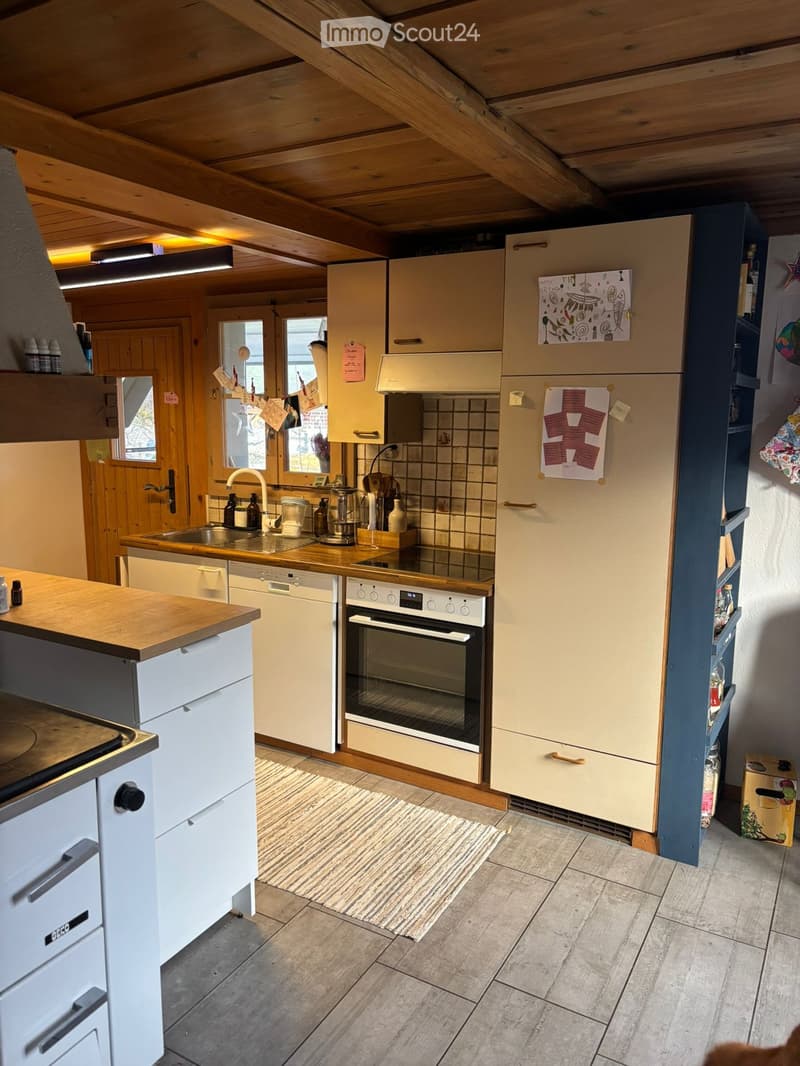 Bright kitchen with wooden ceiling beams, light-colored cabinets, and modern appliances including an oven, refrigerator, and dishwasher. Wooden countertops and tiled floor.