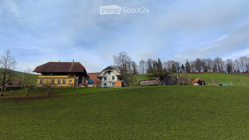 A two-story house with a yellow exterior and a brown roof, situated on a grassy field with trees and hills in the background.