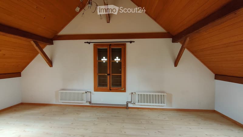 Empty attic room with wooden slanted ceilings, wooden floor, large window with double glass panels, and two radiators on either side of the window.
