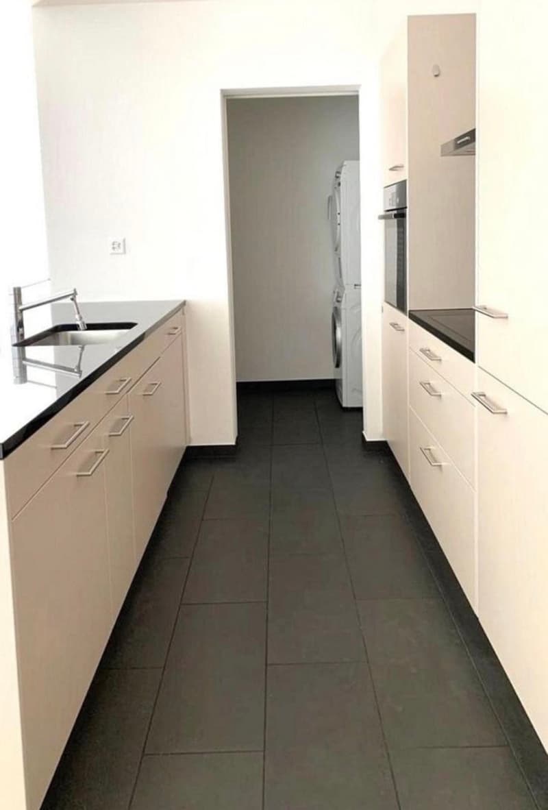 A narrow hallway with white cabinets on both sides, a sink on the left, and a doorway leading to a room with a washing machine and dryer visible.