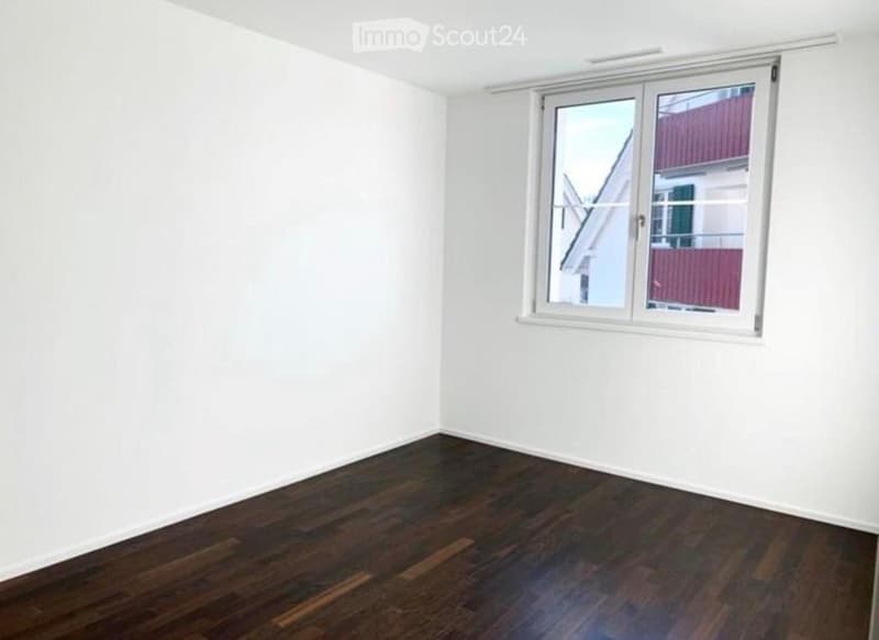 Empty room with white walls and dark wooden flooring, featuring a large window with white frame and blinds.