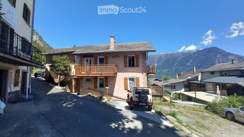 A two-story residential building with a wooden balcony, situated in a picturesque mountain village with clear blue skies and surrounding mountains.