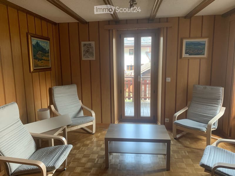 Bright living room with wooden floors, striped walls, and large windows leading to a balcony. Features a sofa, coffee table, and several chairs.