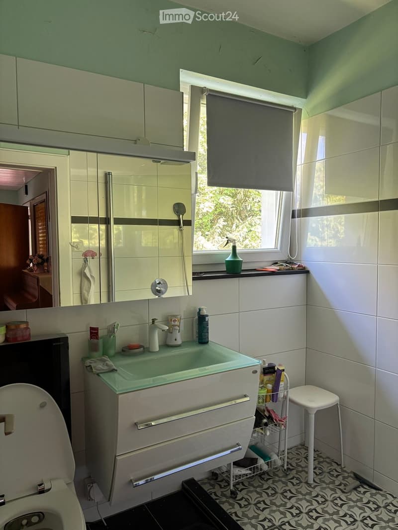 Modern bathroom with a white cabinet, sink, mirror, and toilet. Features a window with a roller shade, a small stool, and various toiletries and cleaning supplies on the windowsill and counter.