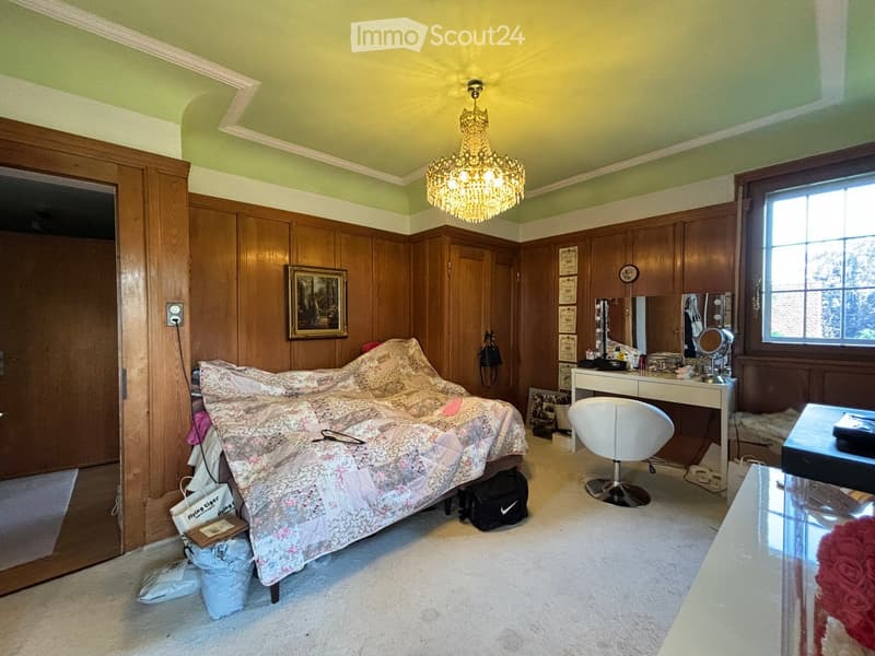 Bedroom with wooden panel walls, a chandelier, a bed with floral bedding, a vanity table with a mirror, a white chair, and a glass table.
