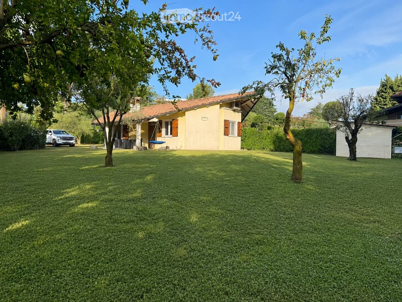A well-maintained lawn with trees and a small house with a tiled roof and shutters in the background.
