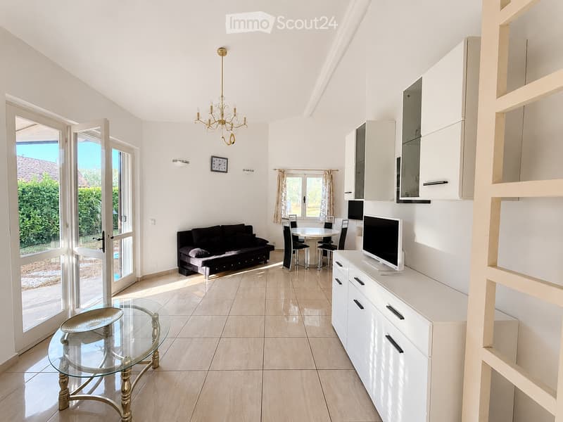 Bright living room with white walls, light wooden flooring, and large windows. Features a black sofa, glass coffee table, dining table with chairs, TV on a white cabinet, and a chandelier.