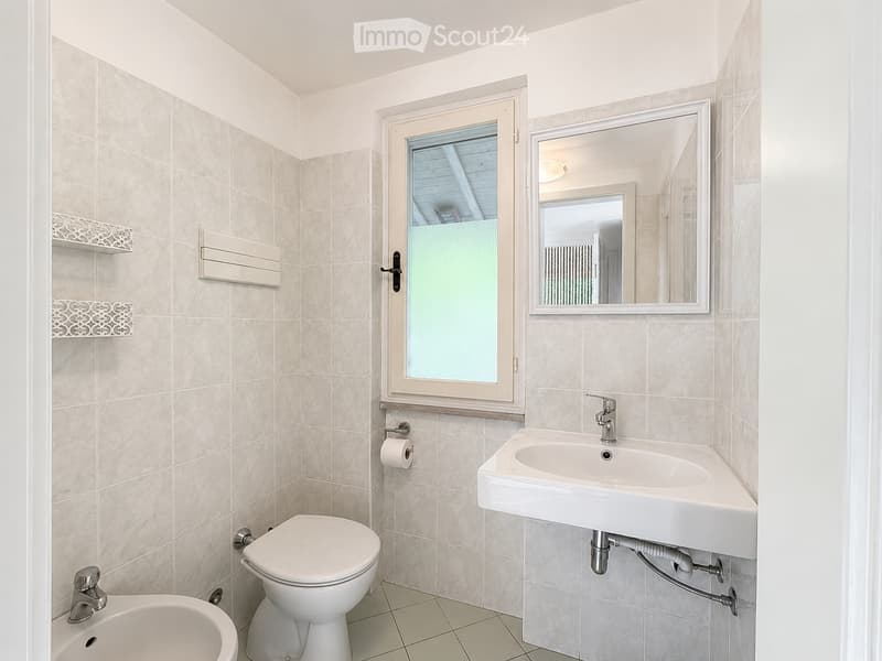 Modern bathroom with beige tiled walls and floor, featuring a toilet, sink with faucet, mirror, and a window with frosted glass.