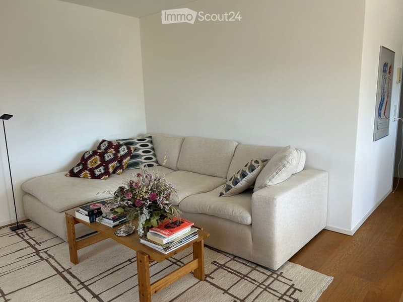 Living room with a beige sectional sofa, wooden coffee table, books, flower arrangement, and a floor lamp.