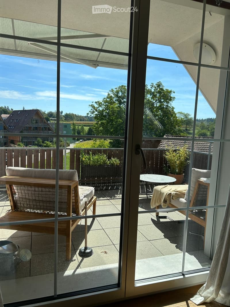 Balcony with a sofa, table, chairs, and plants, overlooking a green landscape with trees and houses.