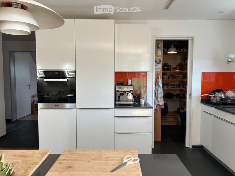 Modern kitchen with white cabinets, stainless steel appliances, and a bright orange backsplash. Features include a refrigerator, oven, coffee machine, and open shelving with jars and bottles.
