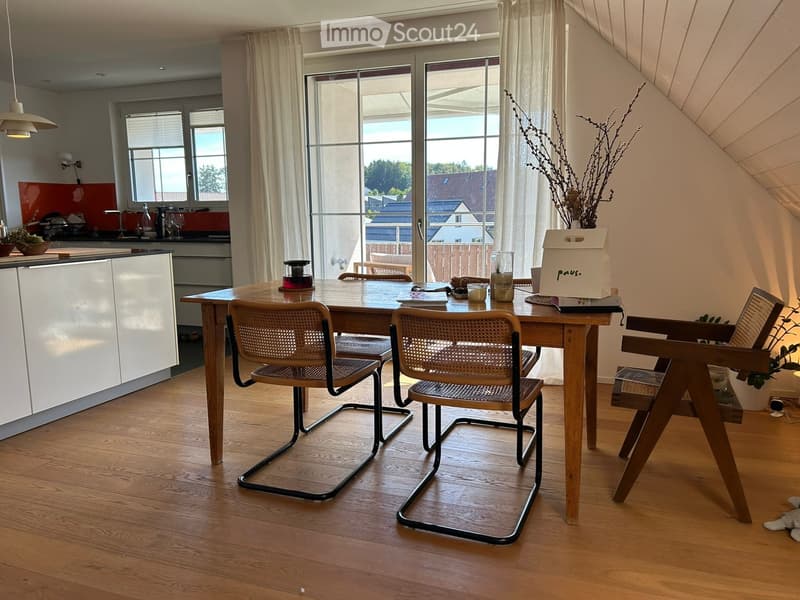 A modern dining area with a wooden dining table, four chairs, and a kitchen area visible in the background. The space features large windows with white curtains, wooden flooring, and a sleek kitchen cabinet.
