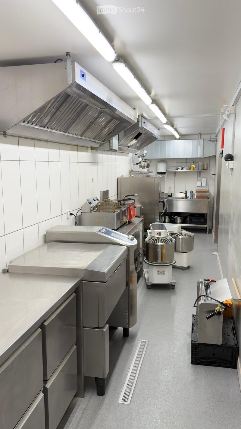 Commercial kitchen with stainless steel appliances, including a refrigerator, mixer, oven, fryer, and sink, arranged around a central counter.