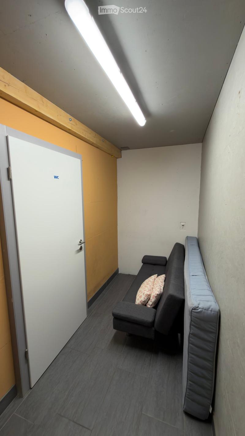 Small, furnished room with a black sofa, two patterned pillows, and a grey mattress leaning against the wall. The room has a slanted ceiling and a white door labeled "WC" on the left side.
