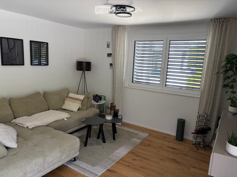 Modern living room with a beige sectional sofa, two coffee tables, a floor lamp, and large windows with blinds. The room features wooden flooring, a white throw blanket on the sofa, and decorative items including a wine rack and potted plants.