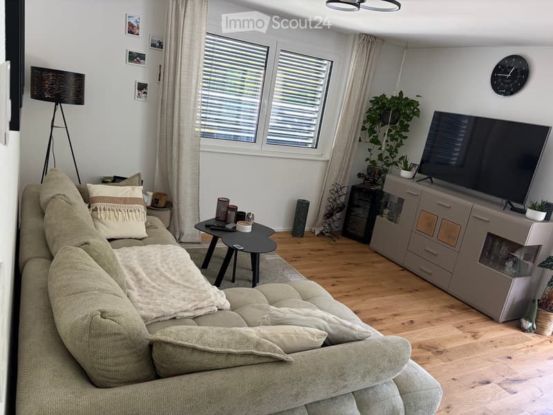 Modern living room with a beige sofa, coffee table, television, side table, floor lamp, clock, and decorative plants.