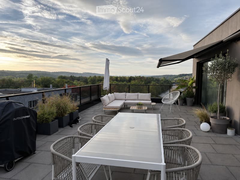 A modern rooftop terrace with a white dining table, wicker chairs, a white sectional sofa, a white hanging chair, potted plants, and a covered grill. The terrace features a panoramic view of a green landscape.