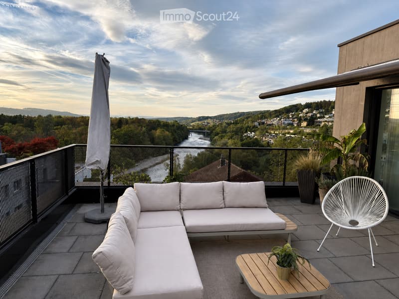 Modern balcony with a white sectional sofa, a white wicker chair, a small wooden table with a potted plant, and an umbrella, overlooking a scenic city view.