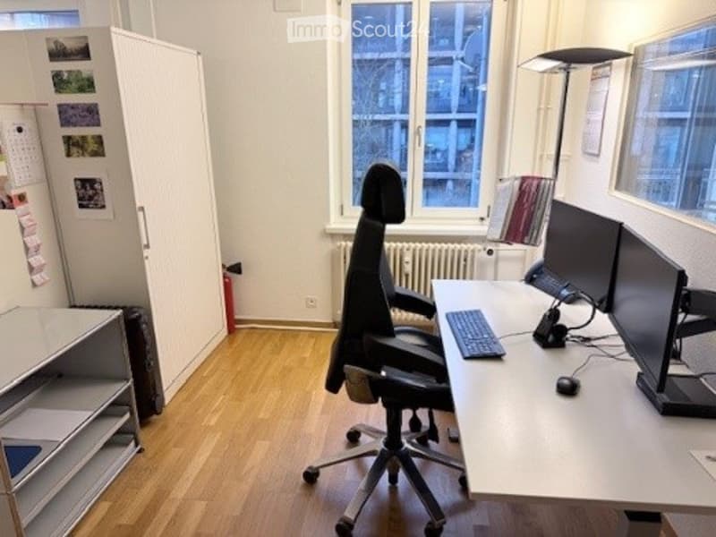 Modern office space with a large desk, ergonomic office chair, dual monitors, keyboard, mouse, and a cabinet with shelves. The room has wooden flooring and large windows offering a city view.