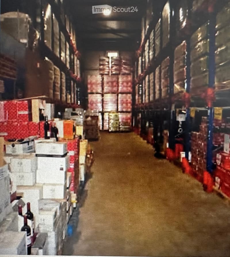 A long, narrow warehouse corridor with metal shelving units on both sides, filled with various boxes and storage items. The floor is concrete, and the ceiling has exposed industrial lighting.