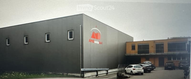 Modern industrial building with a grey facade, logo, and adjacent parking area with several cars.