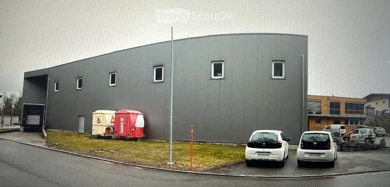 Modern industrial-style building with grey facade, large garage entrance, and several small windows. Two trailers and several cars are parked nearby.