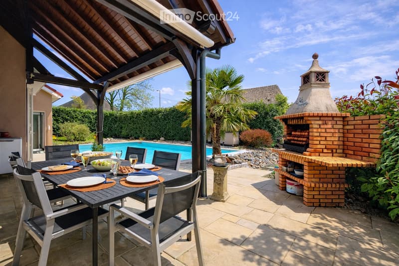 Outdoor patio with a dining table, chairs, and a brick oven. A swimming pool is visible in the background, surrounded by greenery and a covered seating area.