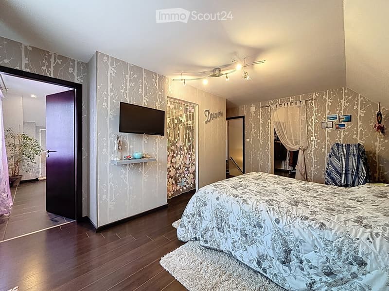Cozy bedroom with wooden flooring, a large bed with floral bedding, a mounted TV, a nightstand with a plant, and decorative picture frames on the wall.