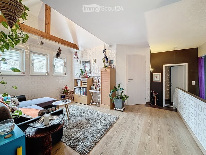 A cozy living room with wooden floors, a patterned rug, a sofa, a coffee table, a bookshelf, and several plants. The room features large windows and a slanted ceiling with exposed wooden beams.