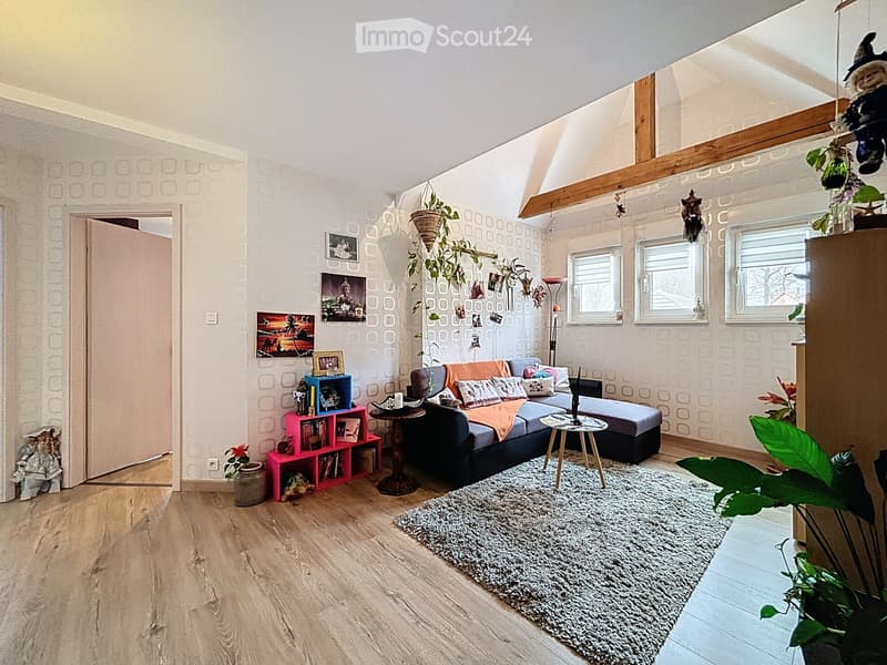 A cozy living room with wooden flooring, a grey sectional sofa, a coffee table, side tables, a shaggy rug, and decorative plants. The walls feature patterned wallpaper and framed artwork.