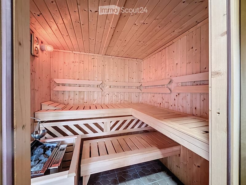 The image shows a wooden sauna with multiple wooden benches at varying heights, a wooden ceiling, and a wooden door. There is a wooden light fixture on the wall, a wooden scale, a wooden bucket, and a wooden stove with rocks inside.