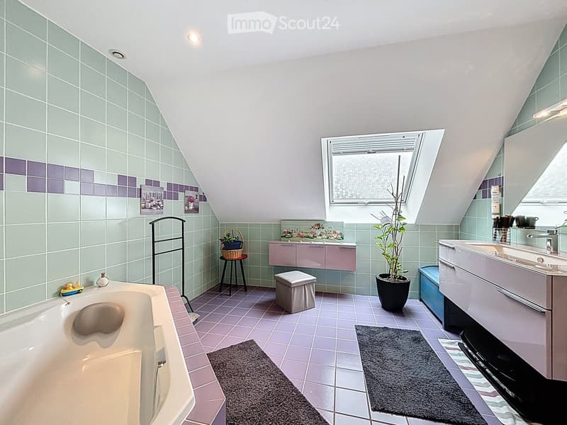 Bathroom with a bathtub, sink, vanity, stool, potted plant, towel rack, and picture frames. The room features light green and purple tiles, a skylight window, and a small window with blinds.