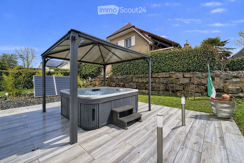 Outdoor patio with a hot tub under a metal canopy, wooden decking, stone wall, and surrounding greenery.