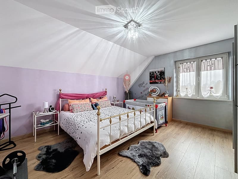 Cozy bedroom with a white metal bed, colorful pillows, wooden nightstand, and decorative items. The room features wooden flooring, a chandelier, and a slanted ceiling with skylight windows.