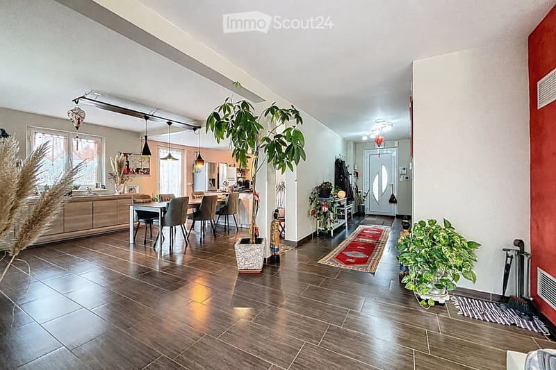 Open-plan living and dining area with wooden flooring, pendant lights, a dining table with chairs, kitchen area with cabinets and refrigerator, and decorative plants.